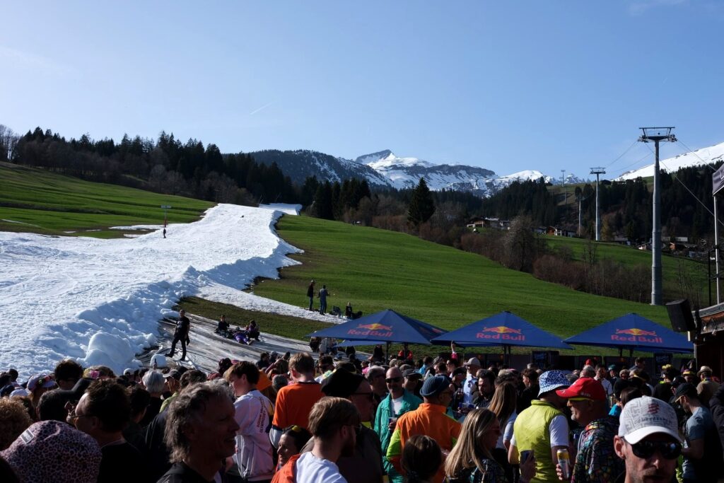 A ribbon of snow running through green grass with a crowd at a spring ski event in Laax