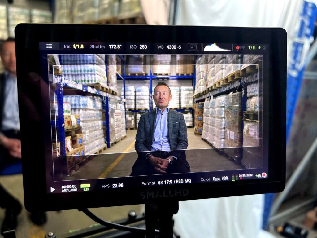 Director monitor showing the Lactalis interview framed among warehouse racking, with frame guides for the ultra-wide screen
