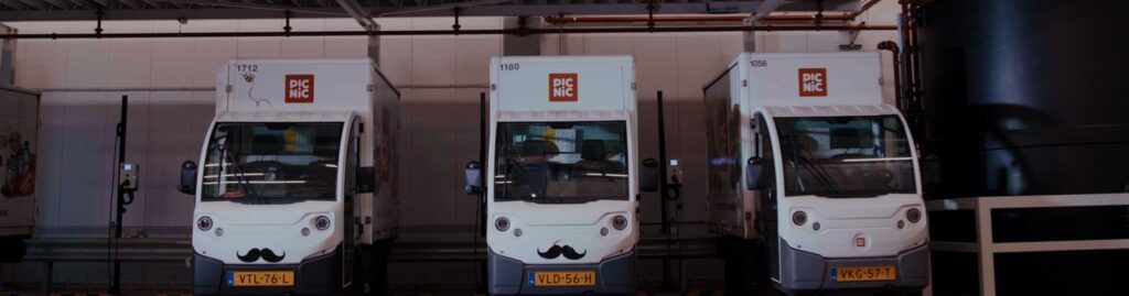 Three Picnic electric delivery trucks in a row, two with moustaches painted on their fronts
